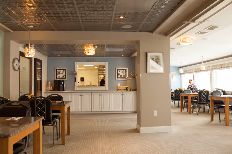 A bright dining area in an assisted living center with several tables and chairs arranged neatly. There is a serving window with a kitchen visible behind it, and a person can be seen working in the kitchen. The room has beige walls, a patterned ceiling, and large windows letting in natural light. A few framed pictures and a clock are on the walls.