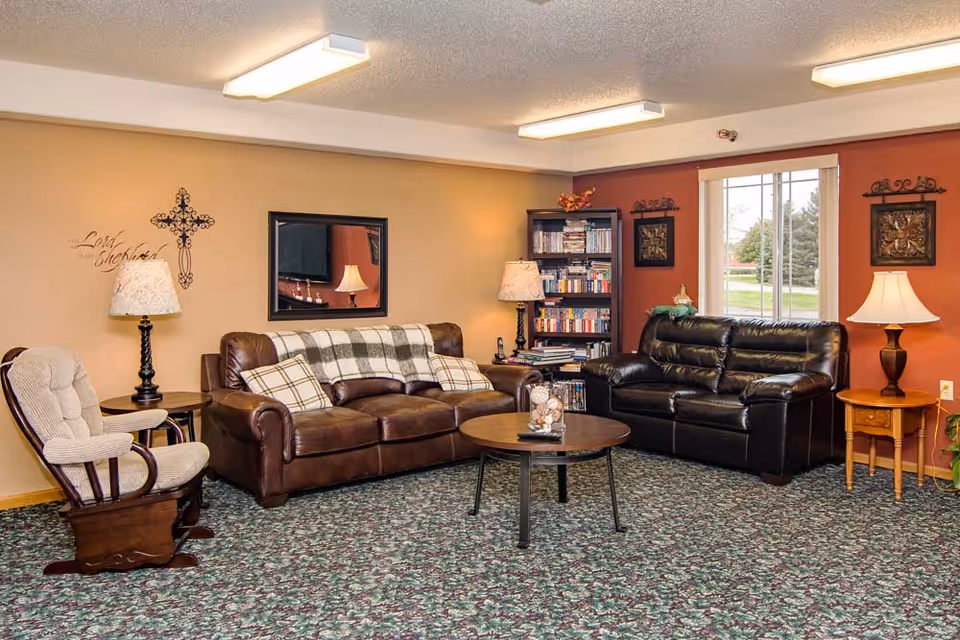 A cozy living room with two leather sofas, one brown and one black, a cushioned rocking chair, two side tables with lamps, a round coffee table, a bookshelf filled with books and DVDs, and a window showing greenery outside. The walls are painted in warm tones with decorative wall art and a mirror.
