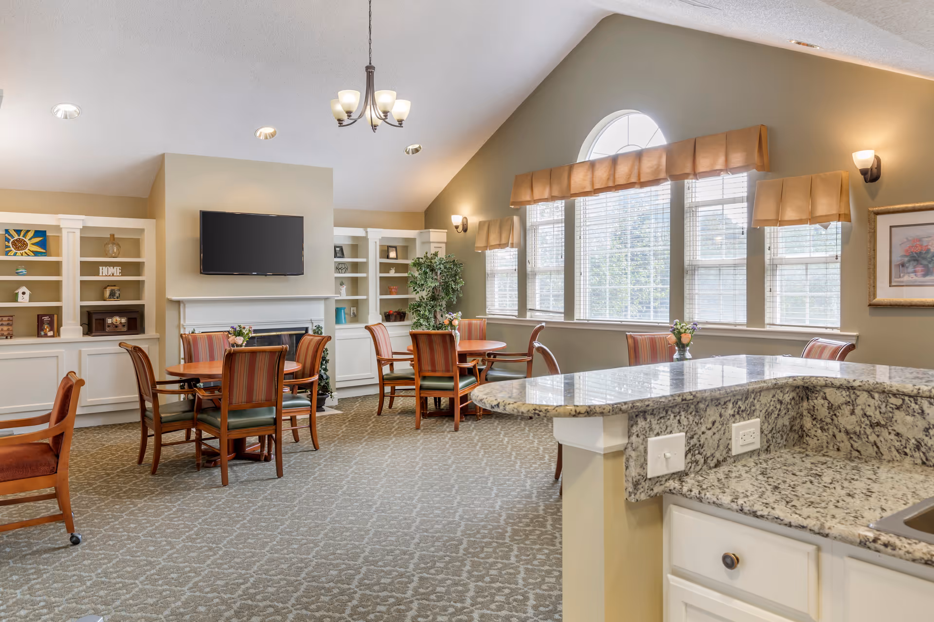 Bright communal dining and lounge area with round tables and chairs, a wall-mounted TV and built-in shelving, and a granite countertop bar in the foreground.