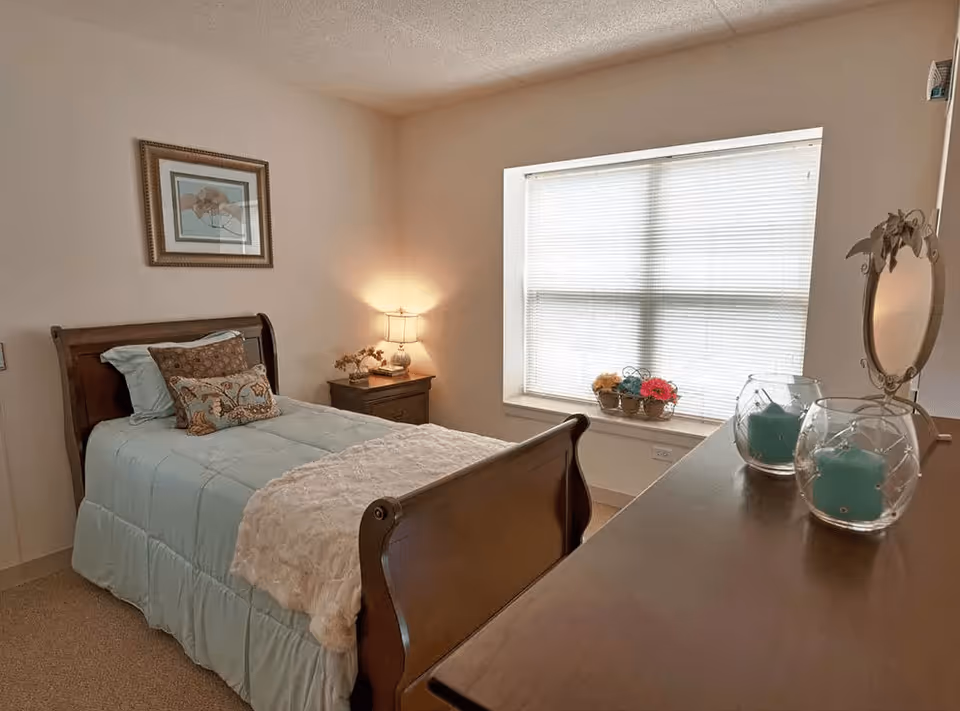 A cozy bedroom with a single wooden sleigh bed made with light blue bedding and decorative pillows. There is a wooden nightstand with a lamp and a small plant next to the bed. A large window with closed blinds lets in natural light, and a wooden dresser with two blue candles in glass holders and a decorative mirror is visible in the foreground.