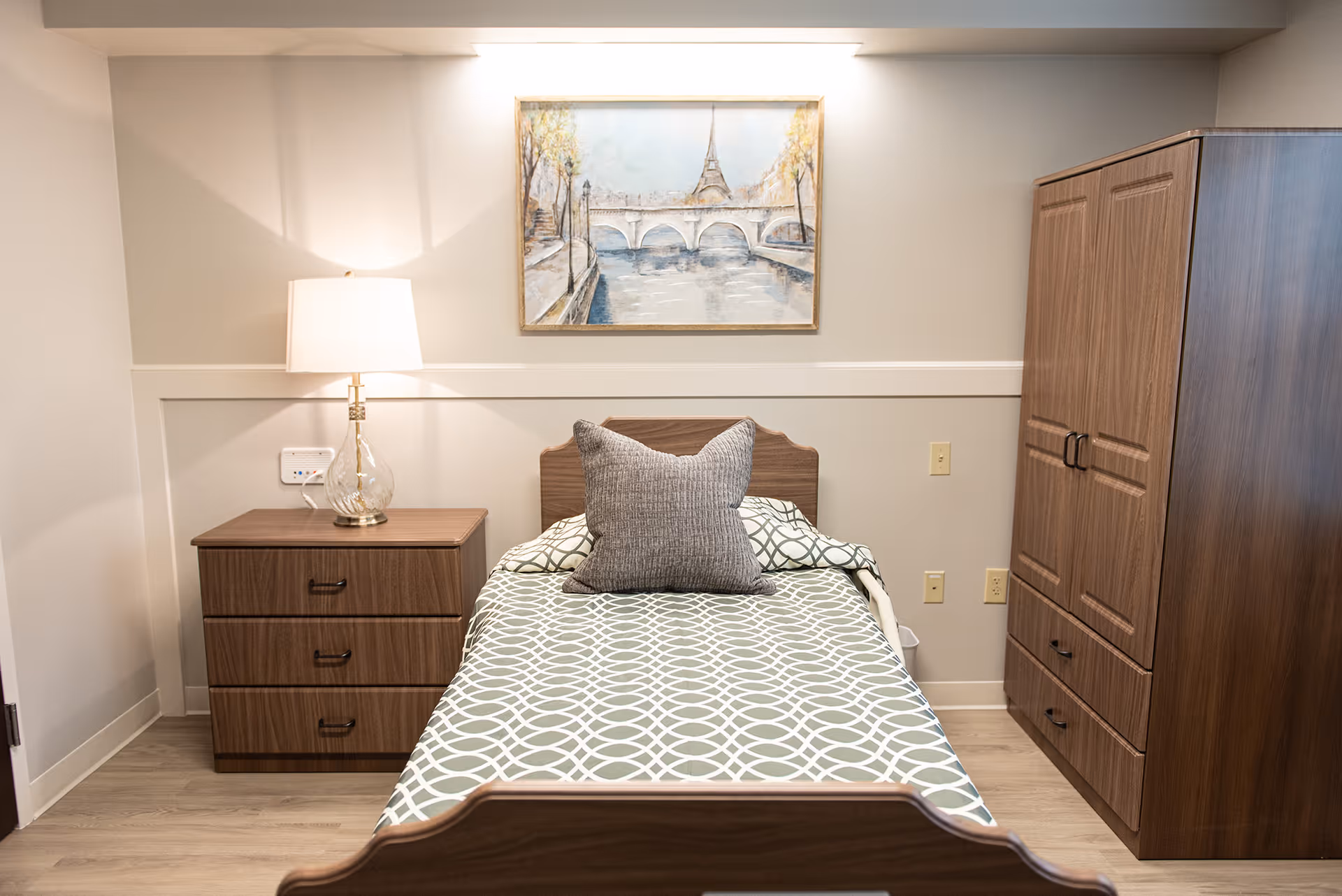 A neatly made single bed with a patterned bedspread and a gray pillow in a room. To the left of the bed is a wooden nightstand with three drawers and a lamp on top. On the right side of the bed is a large wooden wardrobe. Above the bed hangs a framed painting of the Eiffel Tower and a bridge over a river. The room has light-colored walls and wood flooring.
