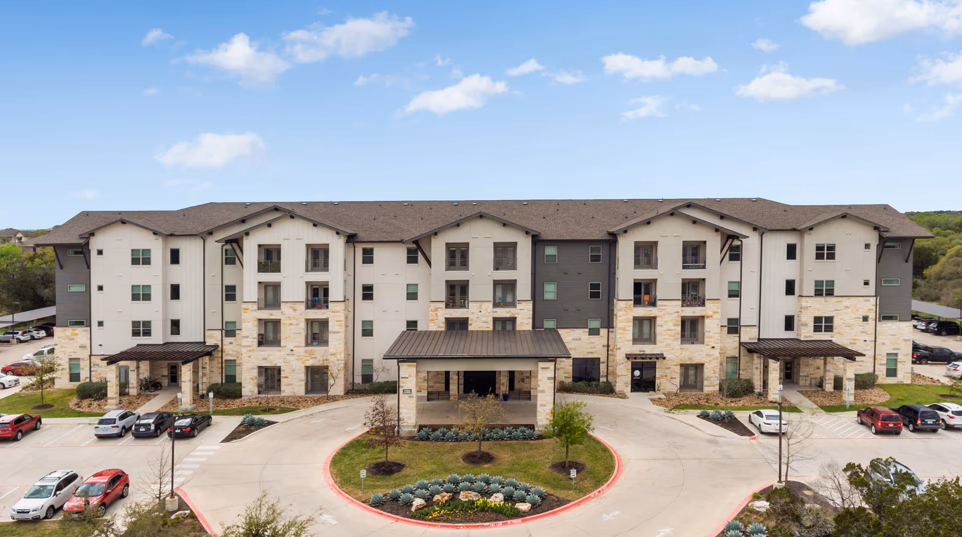 Front view of a four-story stone-and-siding senior living building with covered entry, circular driveway, and parked cars.