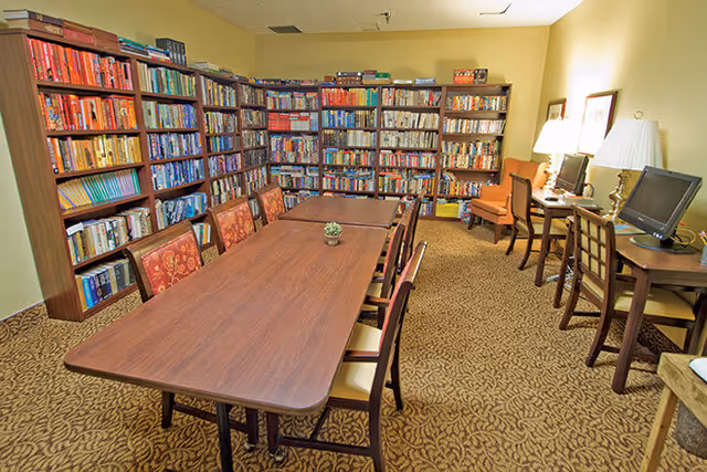 A cozy library room with several tall bookshelves filled with books along the walls. In the center, there is a long wooden table surrounded by chairs with patterned upholstery. On the right side, there are desks with computers and lamps, and a comfortable armchair is placed near the desks. The room has warm yellow walls and patterned carpet flooring.