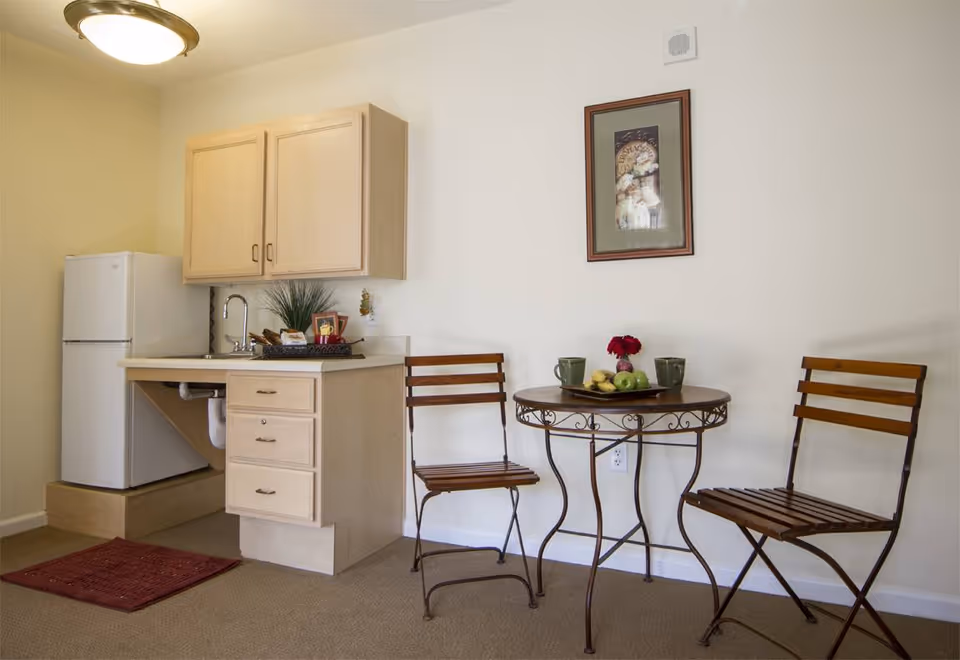 Small kitchenette area with a white refrigerator, light wood cabinets, a sink, and a countertop. Next to the kitchenette is a round metal table with a wooden top, two wooden chairs, two green mugs, a plate with fruit, and a small vase with red flowers. A framed picture hangs on the wall above the table.