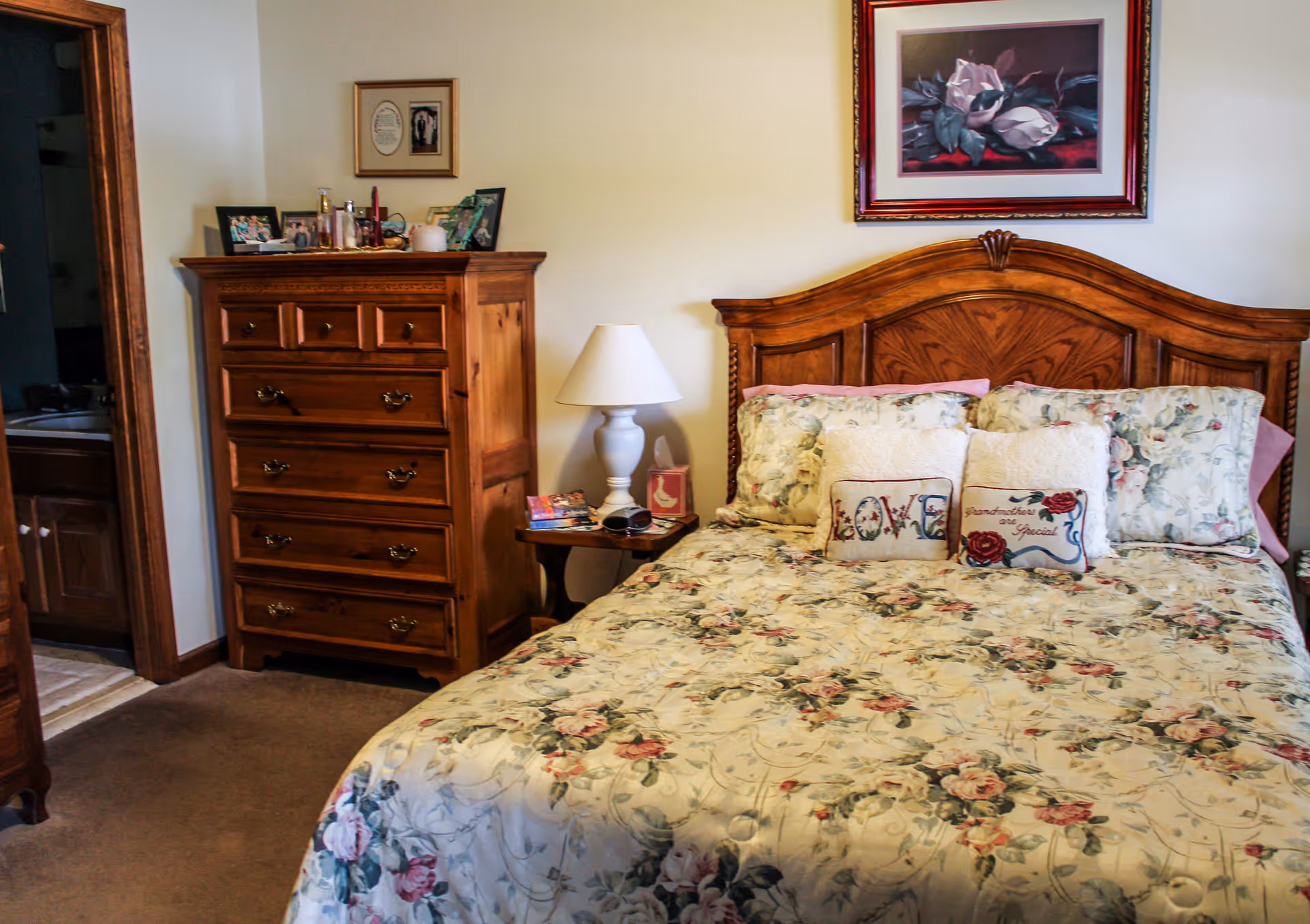 A cozy bedroom featuring a wooden bed with a floral bedspread and decorative pillows, including one that says 'LOVE' and another that says 'Grandmother are Special'. Next to the bed is a wooden nightstand with a white lamp and some books. A wooden dresser with multiple drawers stands against the wall, and a framed floral painting hangs above the bed. An open doorway reveals a glimpse of a bathroom.