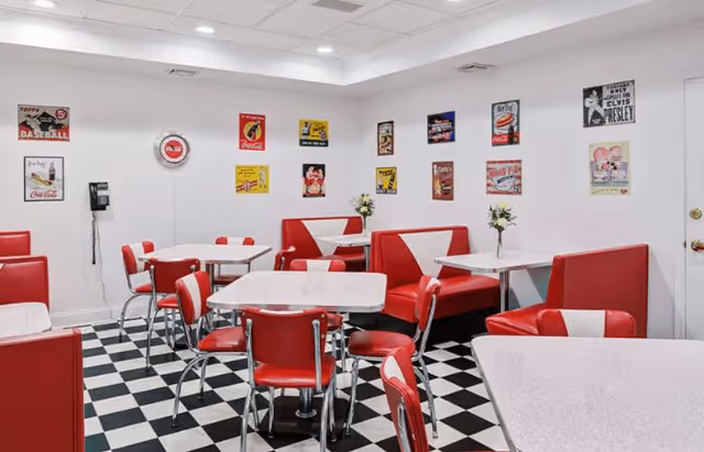 A retro-style dining room with red and white booths and chairs, white tables, and a black and white checkered floor. The walls are decorated with vintage-style posters and signs, including Coca-Cola advertisements and an Elvis Presley poster. A wall-mounted telephone is visible on the left side.
