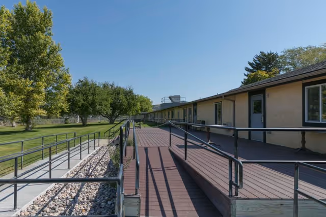 Outdoor view of a single-story building with a wooden ramp and metal railings leading to the entrance. The area is surrounded by green grass, trees, and a clear blue sky.