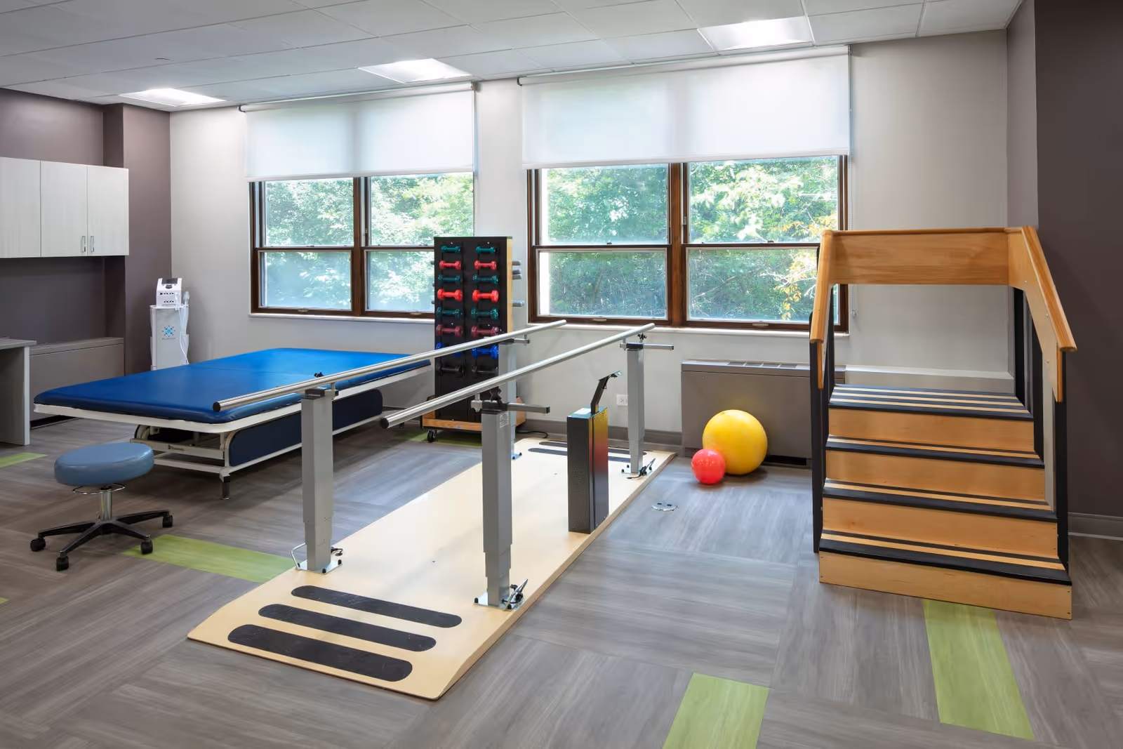 A bright physical therapy room with parallel bars, practice stairs, an exercise table, stability balls and weight rack in front of large windows.
