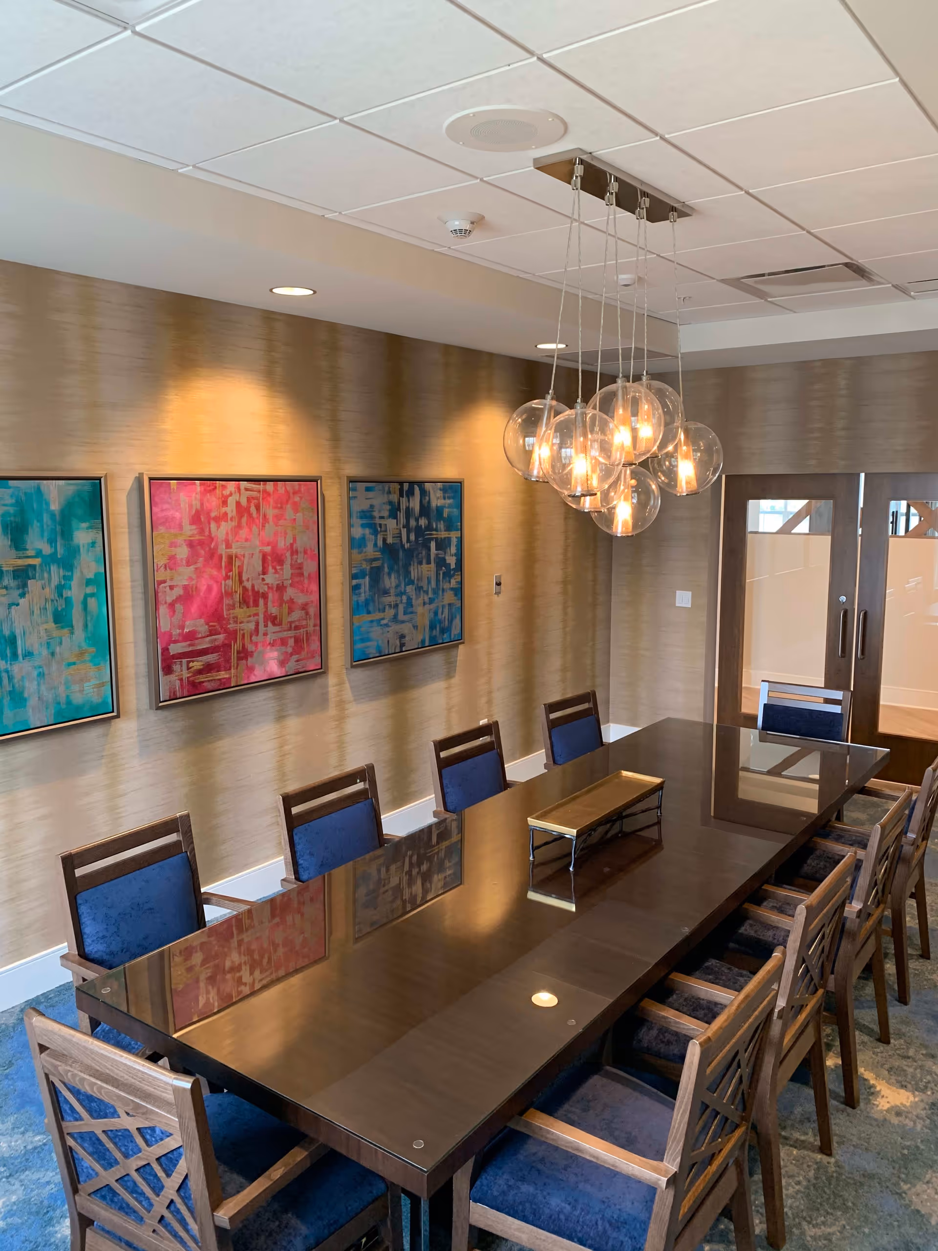 A modern dining room with a long dark wooden table surrounded by twelve wooden chairs with blue cushions. Above the table hangs a contemporary light fixture with multiple glass globes containing light bulbs. The wall behind the table features three colorful abstract paintings in blue, pink, and teal tones. Double wooden doors with frosted glass panels are visible in the background.