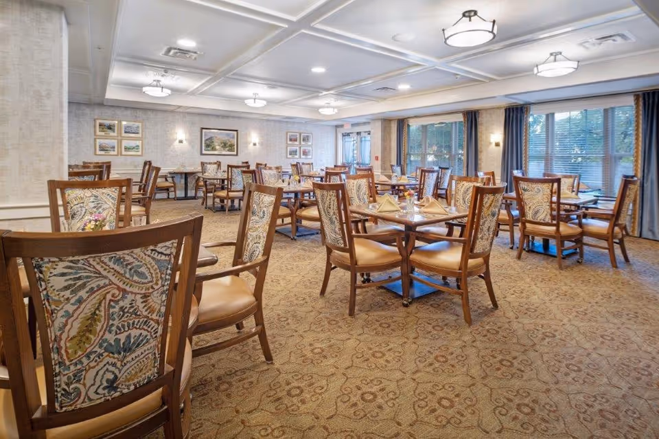 A spacious dining room with multiple wooden tables and chairs arranged neatly. Each table is set with folded napkins and small flower arrangements. The room has large windows with curtains, patterned carpet, framed artwork on the walls, and ceiling lights providing bright illumination.