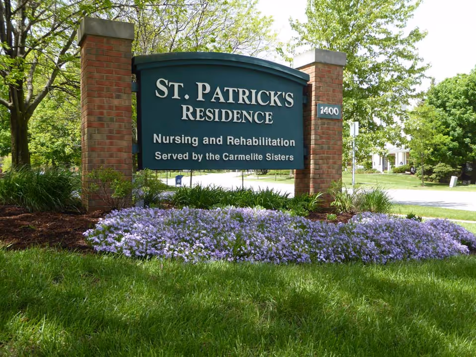 A green sign with white text reading 'St. Patrick's Residence Nursing and Rehabilitation Served by the Carmelite Sisters' mounted between two brick pillars. The sign is surrounded by green grass, purple flowers, and trees in a sunny outdoor setting.