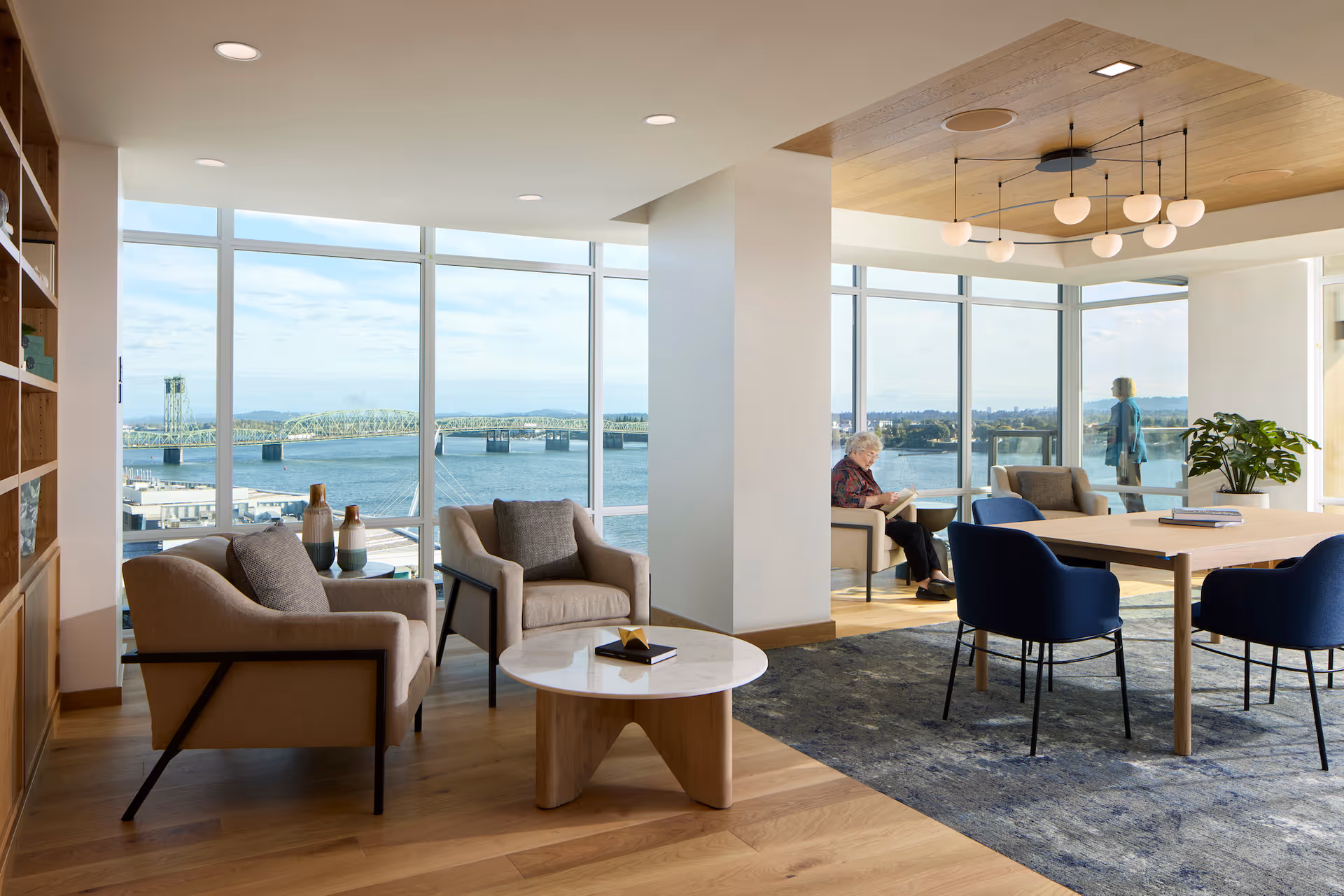 A bright and modern living room area with large floor-to-ceiling windows offering a scenic view of a river and a bridge. The room features comfortable armchairs, a round marble coffee table, a wooden dining table with blue chairs, a bookshelf, and a ceiling light fixture with multiple globe lights. An elderly woman is seated reading a book near the window, and another person is standing by the window looking outside.