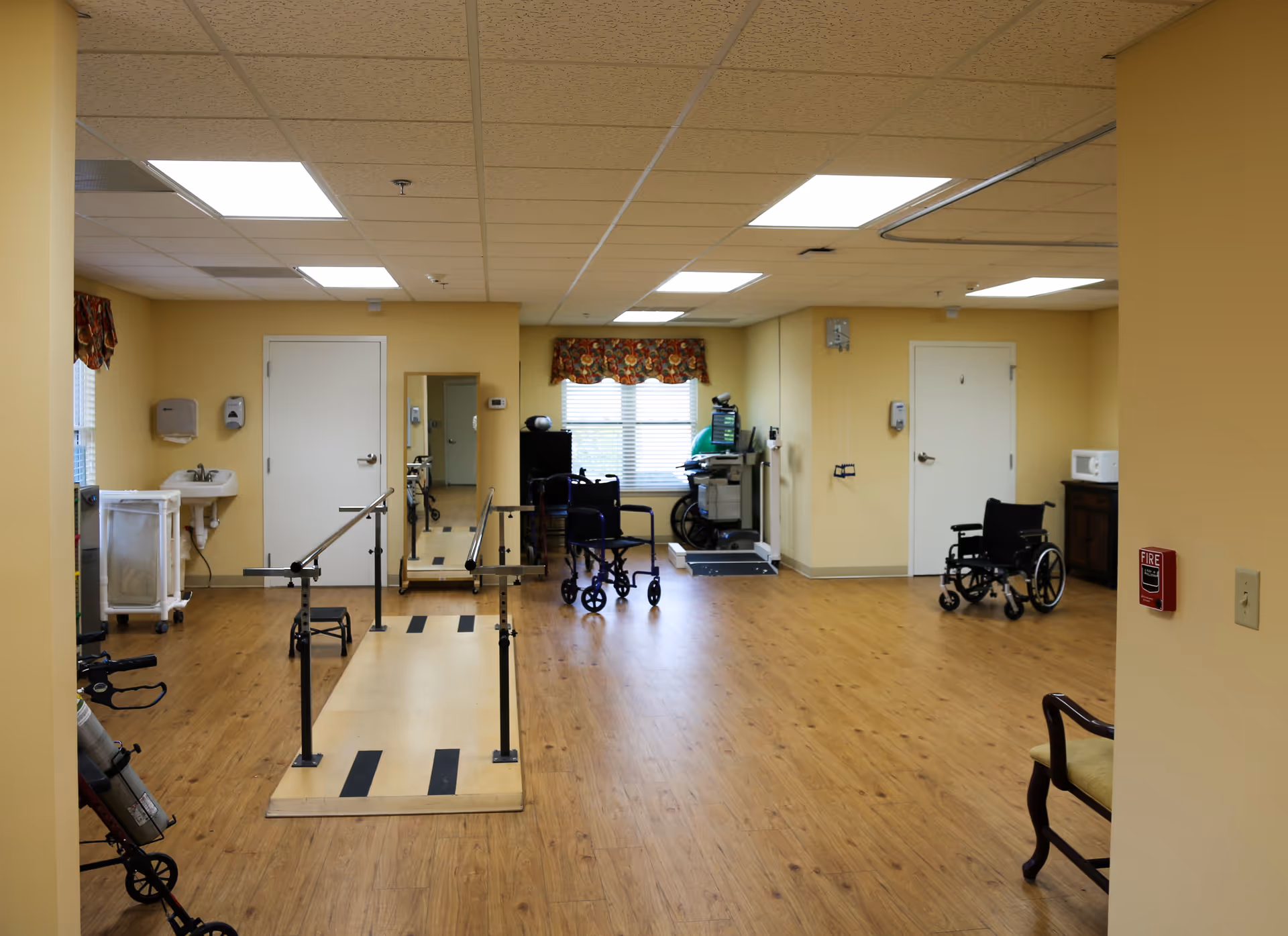 A rehabilitation room with wooden flooring and yellow walls, featuring parallel bars for walking exercises, wheelchairs, a walker, a sink, a mirror, and medical equipment near the windows with floral curtains.