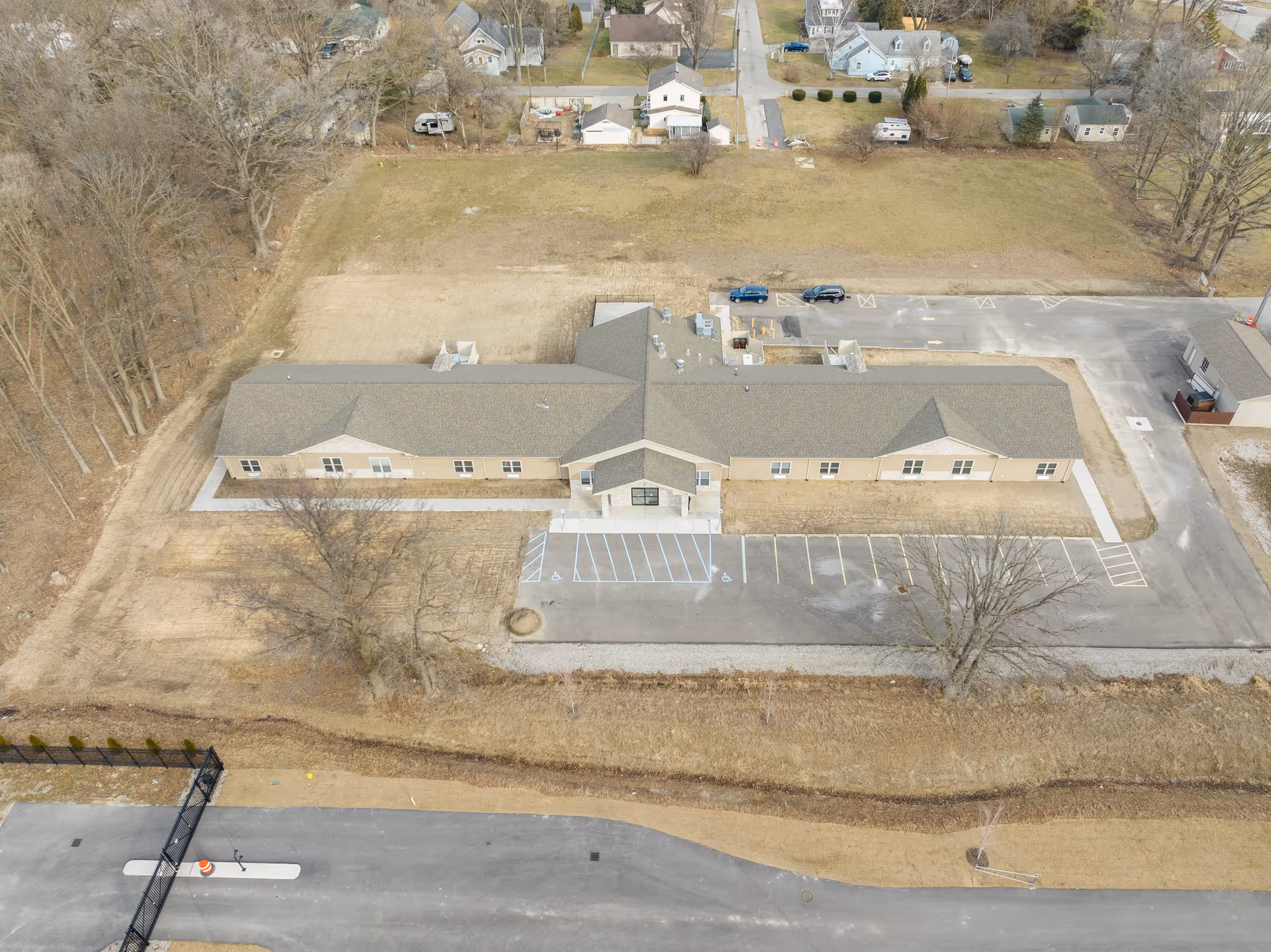 Aerial view of a single-story building with a large parking lot in front, surrounded by open grassy areas and trees. The building has a central entrance with a covered porch and multiple windows along its length. Residential houses and streets are visible in the background.