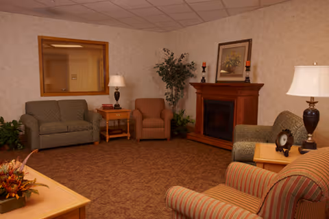 A cozy living room with a green loveseat, a striped armchair, and a brown armchair arranged around a wooden electric fireplace. There are two side tables with lamps and decorative items, a large mirror on one wall, a framed picture above the fireplace, and a potted plant in the corner.