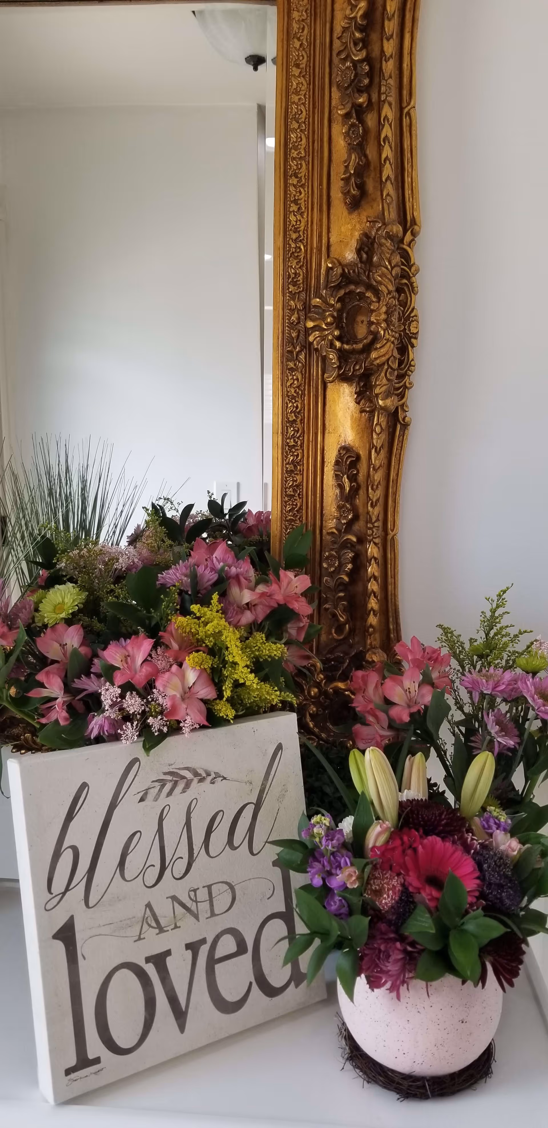 A decorative arrangement featuring a large ornate gold-framed mirror, a bouquet of mixed flowers in various colors including pink, yellow, and purple, and a white sign with the words 'blessed AND loved' written in elegant script.