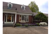 Exterior view of a brick building with a brown roof and dormer windows, featuring a front entrance with glass doors, a bench, and landscaped bushes and flowers in front.