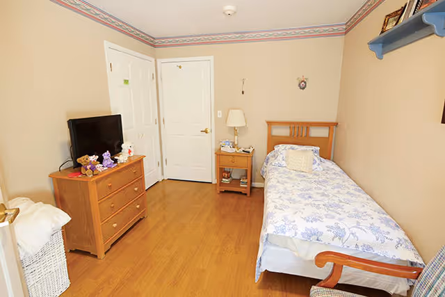 A small bedroom with a single bed covered in a white and blue floral bedspread. Next to the bed is a wooden nightstand with a lamp and some items on it. Across from the bed is a wooden dresser with a TV on top and several stuffed animals. The room has light beige walls with a decorative border near the ceiling and wooden flooring. There are two white doors on the far wall and a wicker laundry basket in the corner.