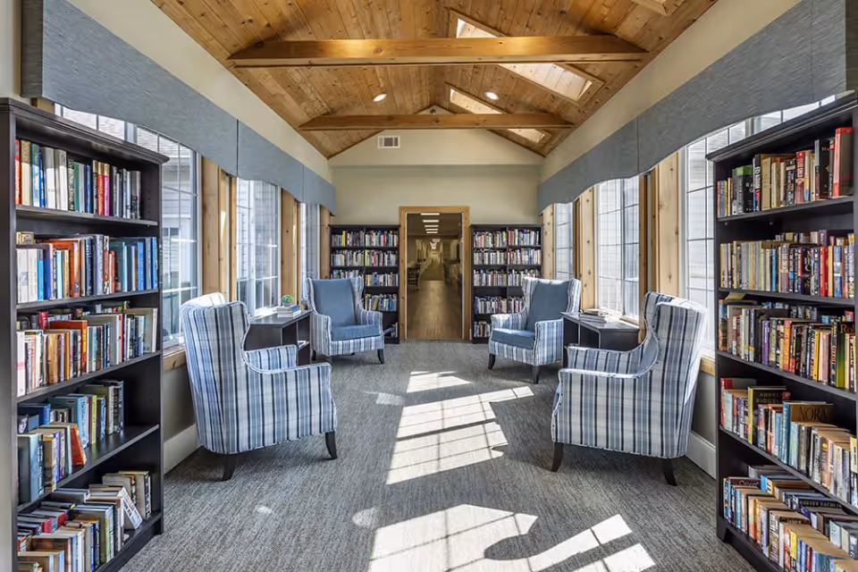 A bright and cozy reading room with large windows on both sides letting in natural light. The room features a wooden vaulted ceiling with exposed beams and skylights. There are four plaid upholstered armchairs arranged in pairs facing each other, with small tables between them. Bookshelves filled with books line both sides of the room and the far end, creating a welcoming library atmosphere.