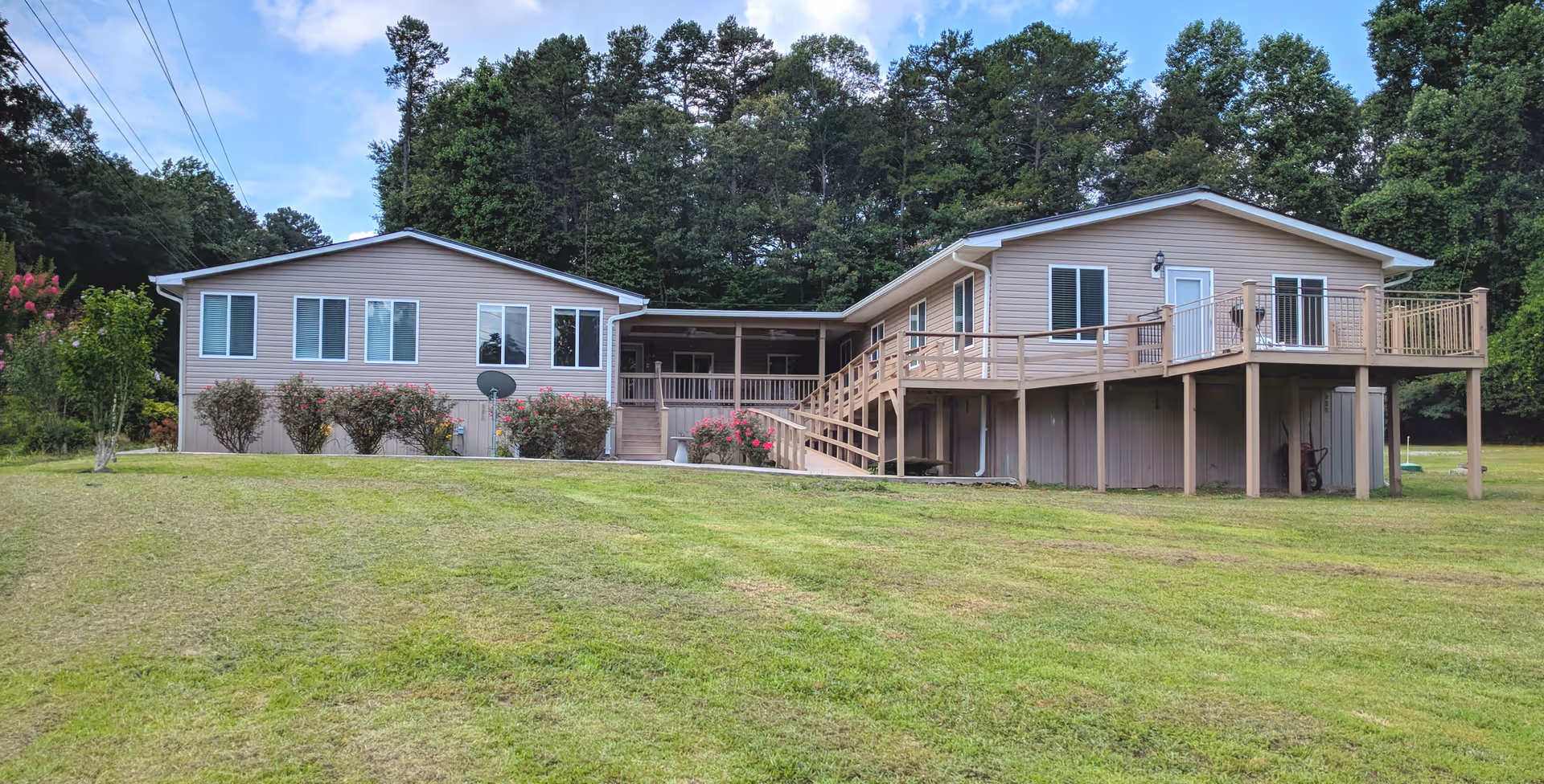 Exterior view of a single-story residential building with beige siding, multiple windows, a raised wooden deck with railings, and stairs leading down to a grassy lawn. The building is surrounded by trees and bushes under a partly cloudy sky.