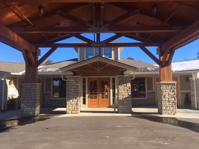 Entrance of a building with a covered driveway supported by wooden beams and stone pillars. The building has double wooden doors decorated with wreaths and windows on either side. The sky is clear and blue.