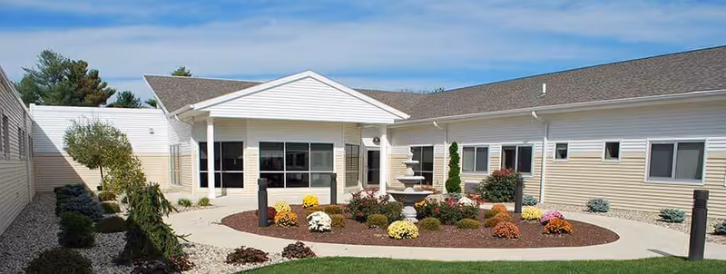Outdoor courtyard area of a senior living facility with a central fountain surrounded by colorful flowers and shrubs, bordered by beige buildings with multiple windows under a blue sky.