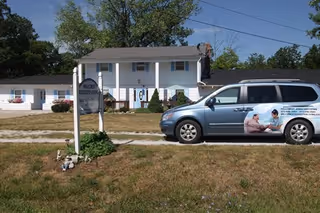 Exterior view of Hillcrest Assisted Living facility showing a two-story white building with a front porch and a sign in the foreground. A silver van with an advertisement for Hillcrest Assisted Living is parked on the grass near the building. Trees and a clear sky are visible in the background.