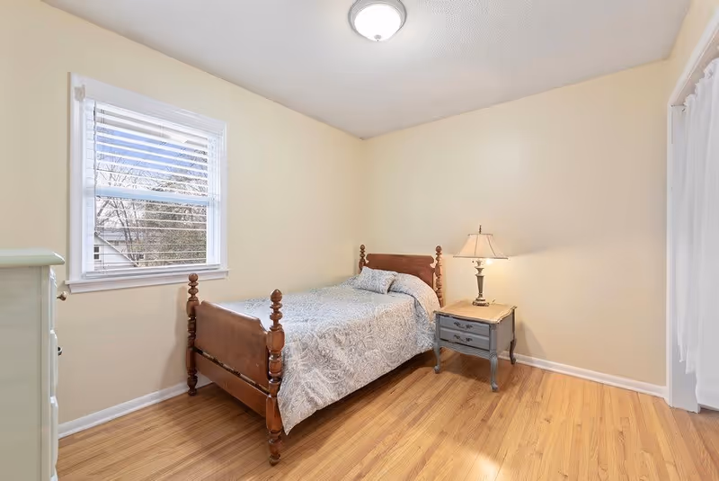 A simple bedroom with a wooden bed frame, a patterned bedspread, a small bedside table with a lamp, a window with white blinds, and light-colored walls and hardwood flooring.