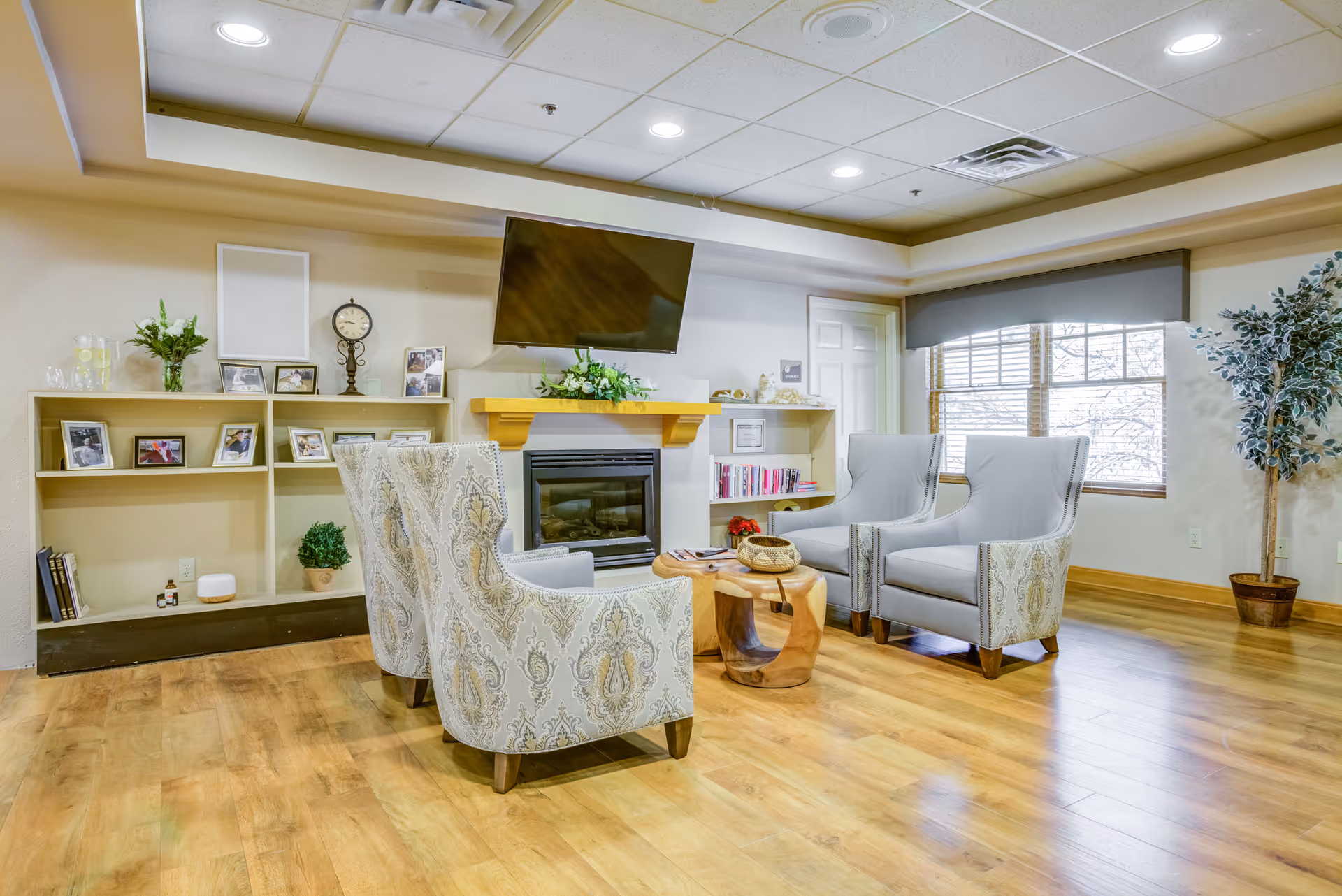 A cozy senior living community lounge area with four upholstered armchairs arranged around a small wooden coffee table. The room features a wall-mounted flat-screen TV above a fireplace, built-in shelves with framed photos, books, and decorative items, a large window with blinds, and a potted plant in the corner. The space has wood flooring and a drop ceiling with recessed lighting.