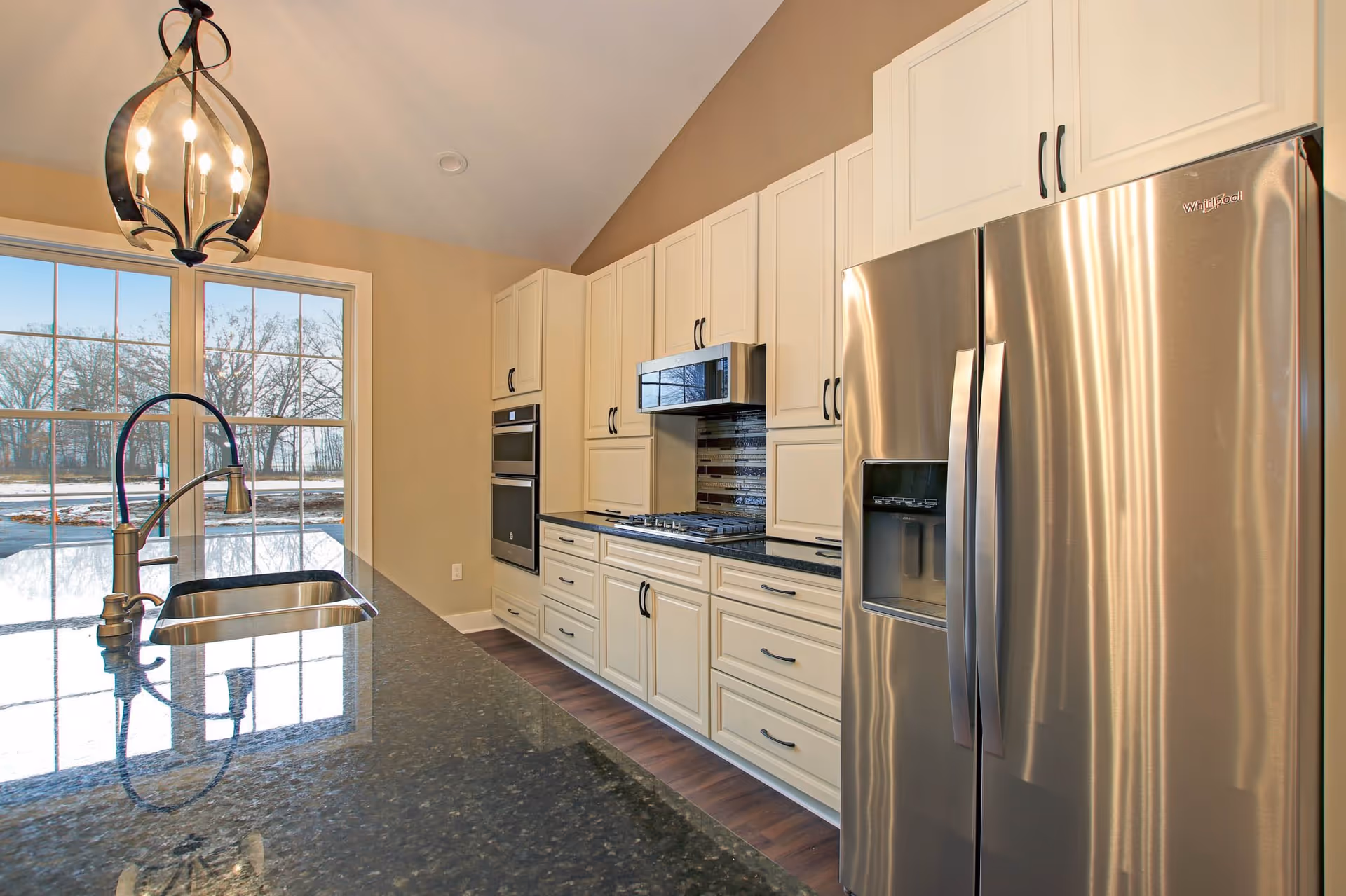 Modern kitchen interior with cream-colored cabinets, a stainless steel refrigerator, built-in oven, microwave, and a black granite countertop island with a sink and faucet. Large window in the background shows an outdoor view with trees and snow on the ground.