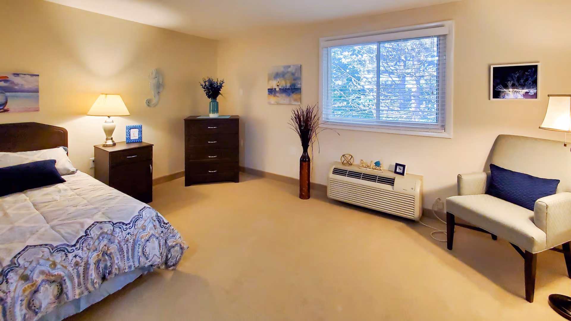 A cozy bedroom with a bed on the left side covered with a patterned comforter and pillows. Next to the bed is a nightstand with a lamp and a small framed picture. A dark wooden dresser with a vase of flowers is against the far wall. A window with blinds lets in natural light above a heating/cooling unit decorated with small ornaments. On the right side, there is a cushioned armchair with a dark blue pillow and a floor lamp beside it. The walls are decorated with framed artwork and a seahorse wall hanging.