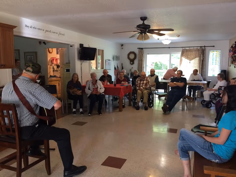 A group of elderly people seated in a common room of an assisted living facility, listening to a man playing guitar. The room has a ceiling fan, a wall clock, a television, and tables with red and striped tablecloths. There is a large window with curtains letting in natural light.