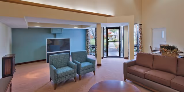 A spacious living room area with two patterned armchairs and a brown sofa arranged around a round wooden coffee table. There is a large flat-screen TV mounted on a blue accent wall, and glass doors leading outside, allowing natural light to enter. The room has beige walls and carpeted flooring.