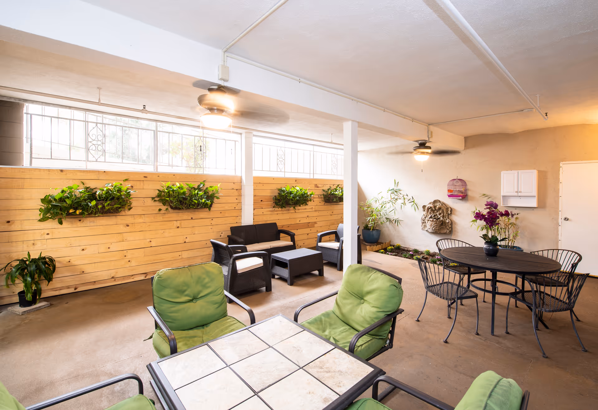 Indoor seating area with green cushioned chairs around a tiled table in the foreground, black wicker chairs and a small table against a wooden wall with mounted plants, and a round black metal table with matching chairs and flower pots near a beige wall with decorative elements and a door. Ceiling fans with lights are mounted on the ceiling.