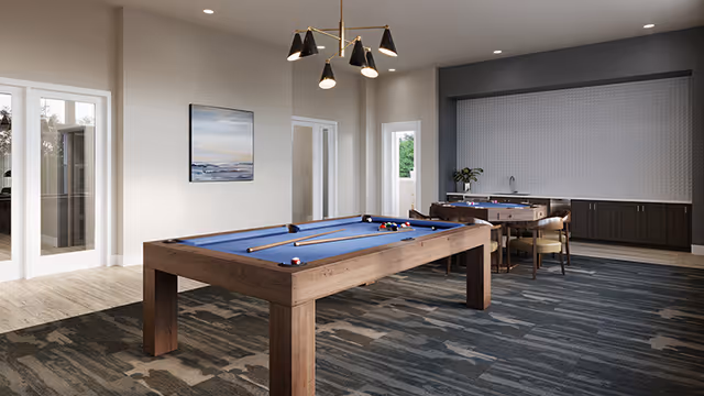 A modern game room with a blue felt pool table in the center, pool cues resting on it, and a foosball table with chairs in the background. The room has a patterned carpet, neutral-colored walls, a contemporary chandelier, and a small kitchenette area with cabinets and a sink.
