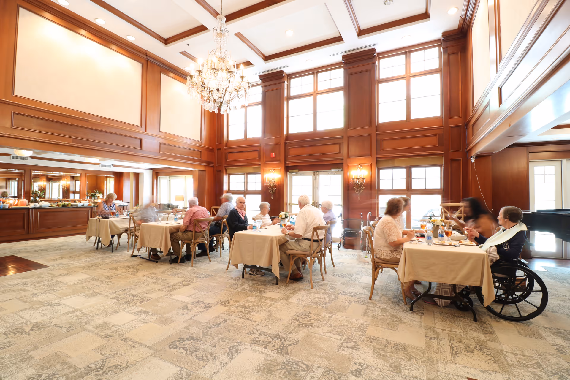 A bright dining room with high ceilings and large windows. Several elderly people are seated at tables covered with beige tablecloths, enjoying meals and conversation. The room features wood paneling, a chandelier, and a carpeted floor.