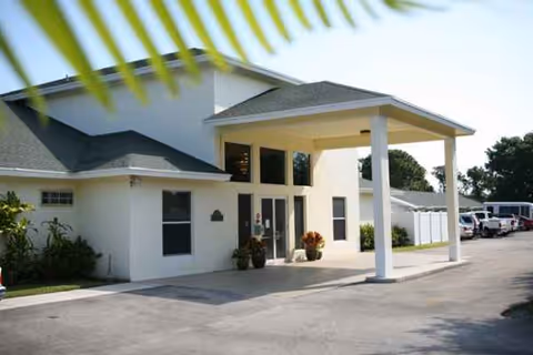 White assisted living facility exterior with a covered entrance and parked cars along the driveway.