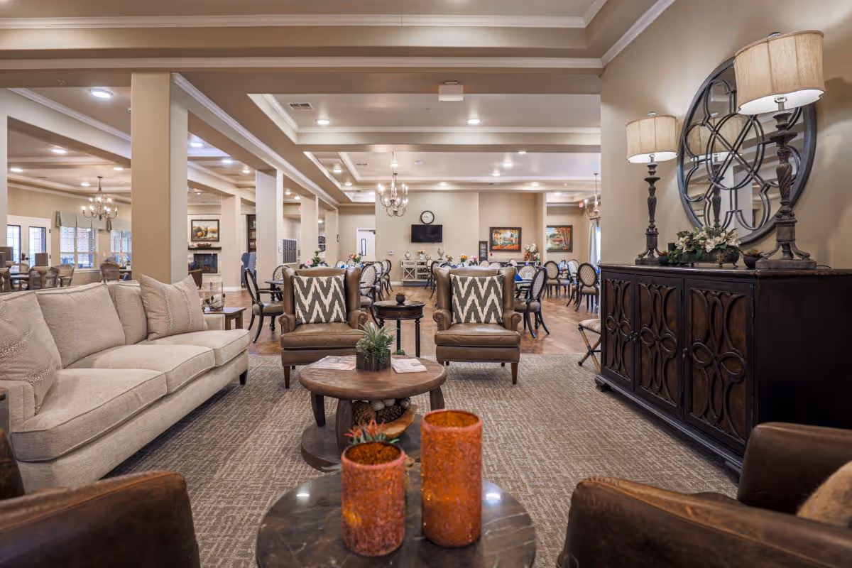 A spacious and well-lit common area in an assisted living facility featuring a beige sofa, two brown armchairs with patterned cushions, a round wooden coffee table with decorative items, and a dark wooden sideboard with two lamps and a large decorative wall clock above it. In the background, there are multiple dining tables and chairs, chandeliers, and framed artwork on the walls.
