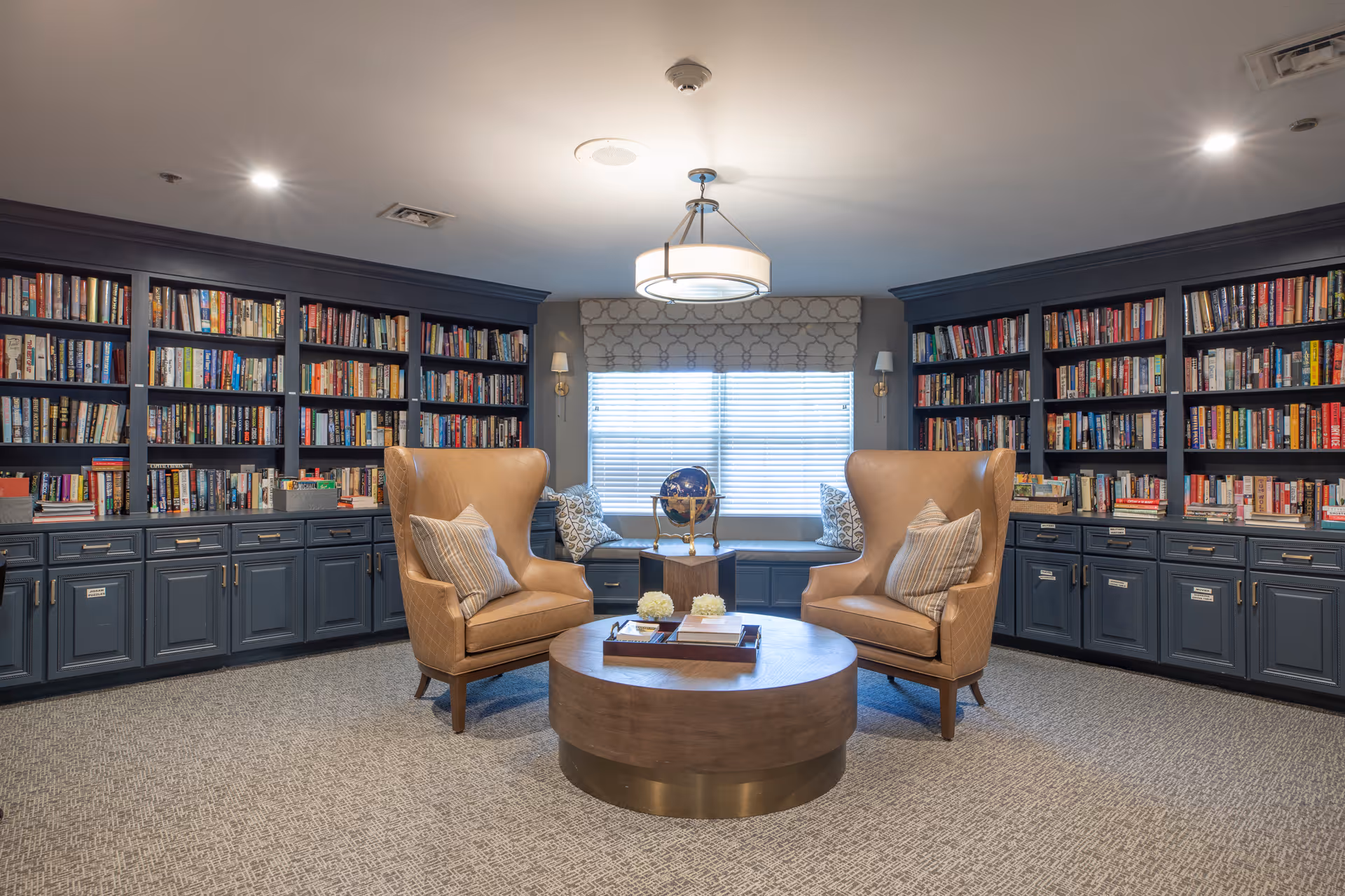 A cozy library room with dark blue built-in bookshelves filled with books lining the walls. Two tan leather wingback chairs with striped pillows face a round wooden coffee table with decorative items on it. A window with blinds and patterned valance is centered on the back wall, flanked by two wall sconces. The room has a carpeted floor and a modern ceiling light fixture.