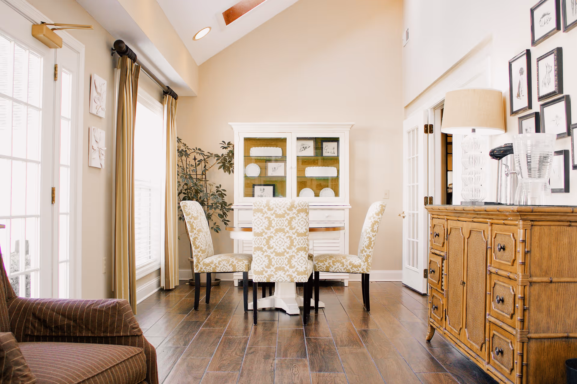A bright dining room with a round wooden table surrounded by four upholstered chairs with a white and beige pattern. Behind the table is a white cabinet displaying plates and framed artwork. To the right is a wooden sideboard with a lamp, water pitcher, and glasses. The room has wooden flooring, beige walls, large windows with beige curtains, and a glass door on the left.
