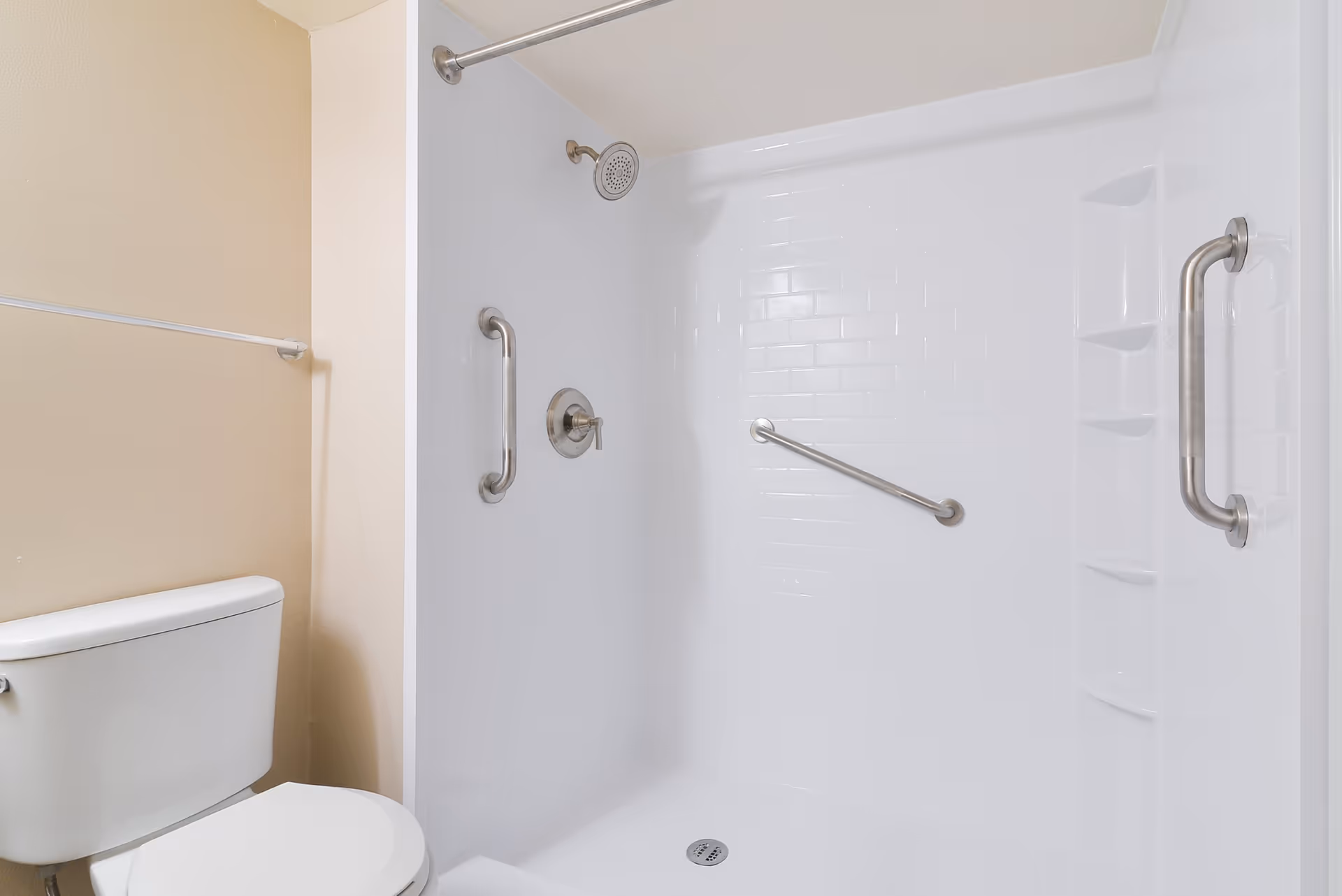 A clean bathroom featuring a white toilet and a walk-in shower with white tiled walls. The shower has multiple stainless steel grab bars and built-in shelves for toiletries.