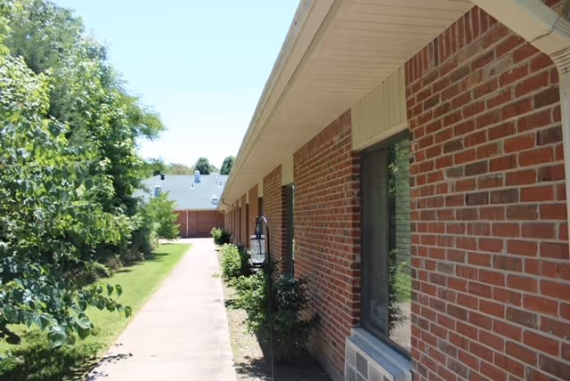 Side view of a brick building with several windows and an air conditioning unit beneath one window. A concrete walkway runs alongside the building, bordered by green grass and trees on the left side under a clear blue sky.