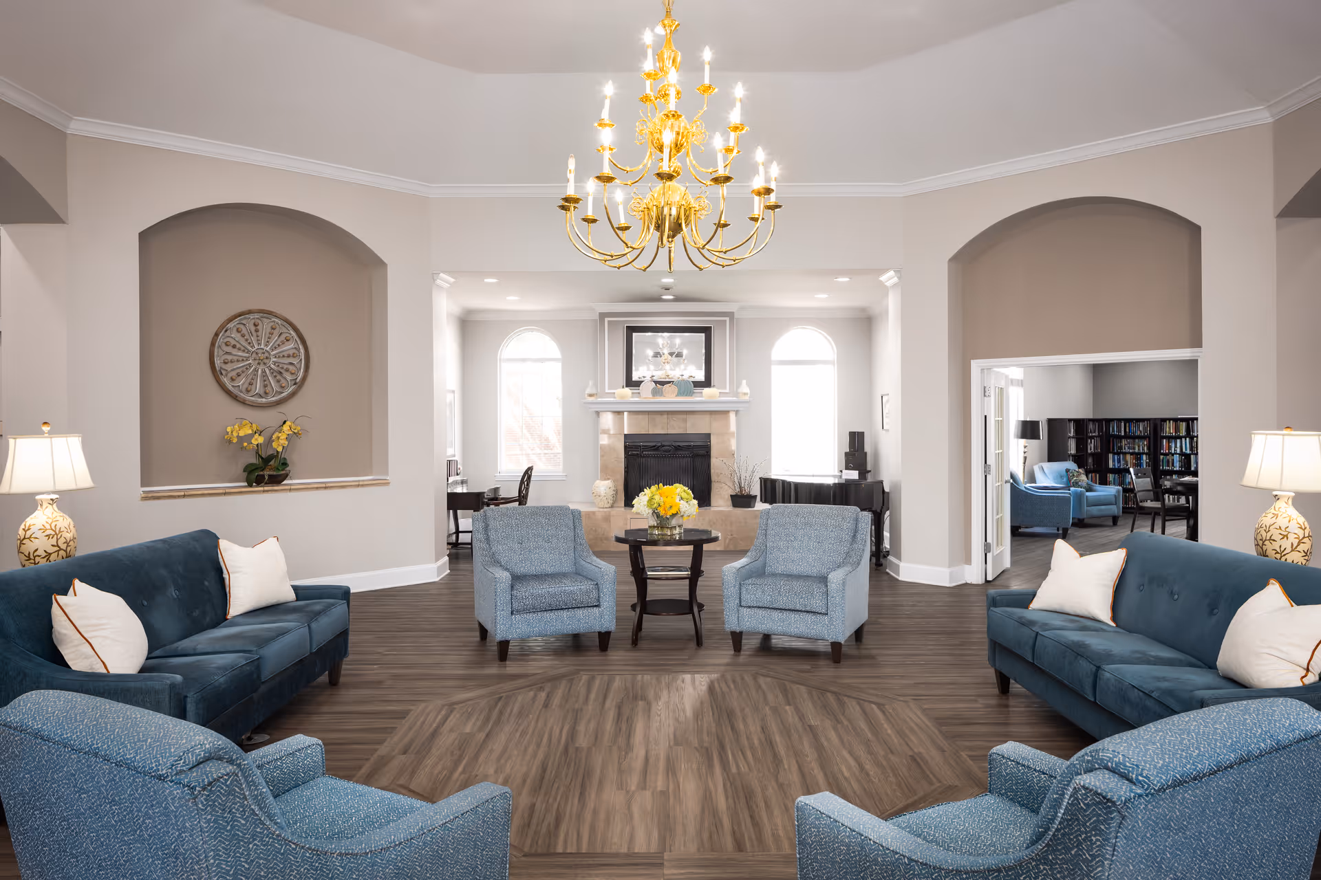 A spacious and elegant living room with a large gold chandelier hanging from the ceiling. The room features two blue sofas and four blue armchairs arranged around a central wooden coffee table with a vase of yellow flowers. In the background, there is a fireplace flanked by two tall arched windows, and a piano is visible to the right. The walls are painted in neutral tones, and the floor is wood with a decorative pattern in the center. There are also table lamps on either side of the sofas and a decorative wall piece with yellow flowers on a shelf to the left.