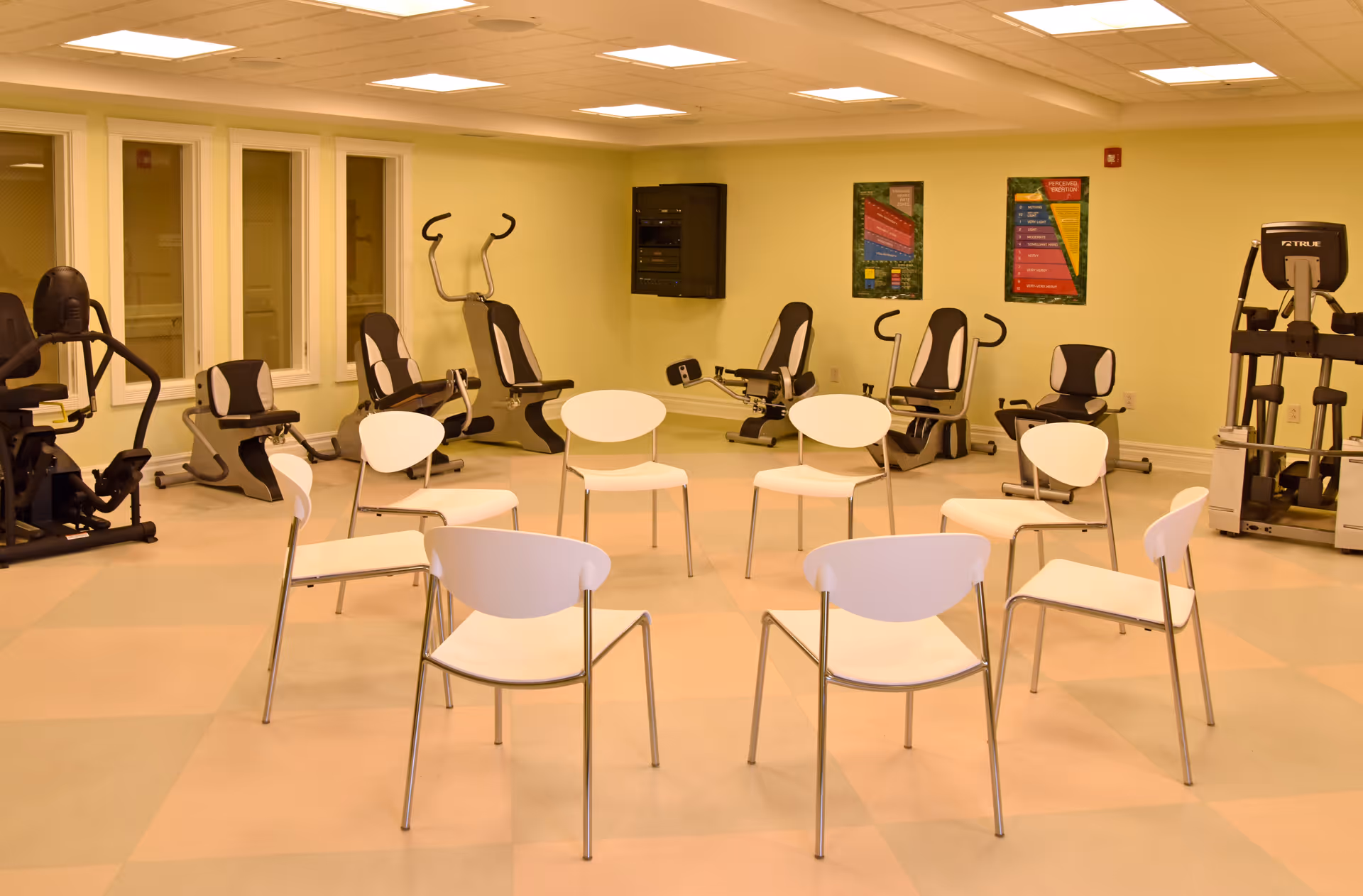 A bright exercise room with white chairs arranged in a circle and several recumbent exercise machines along the walls.