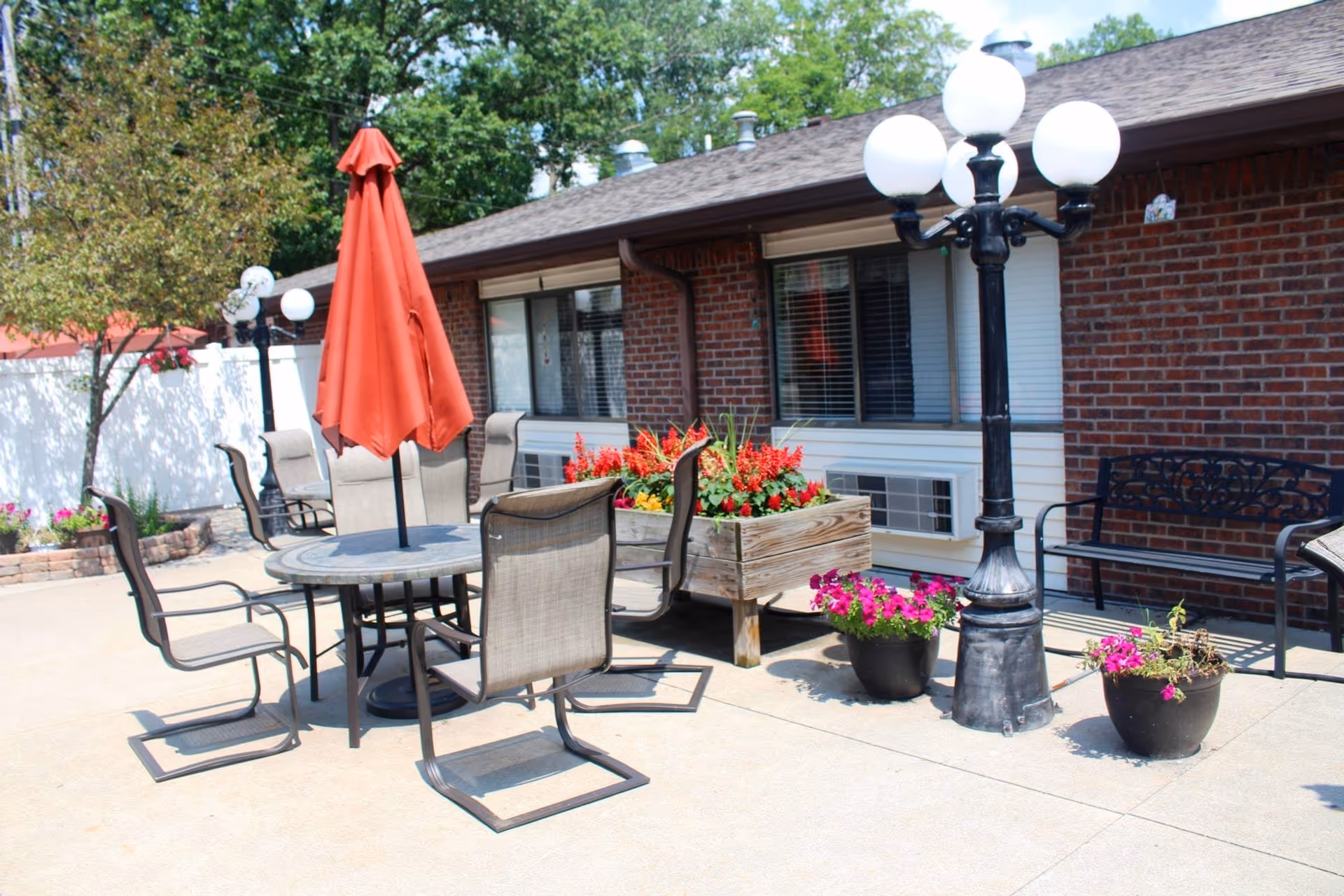 Outdoor patio area with a round table and six chairs, a closed red umbrella in the center of the table, a wooden planter box with red flowers, two black flower pots with pink flowers, a black metal bench, and a vintage-style street lamp. The background shows a brick building with windows and a white fence with trees.