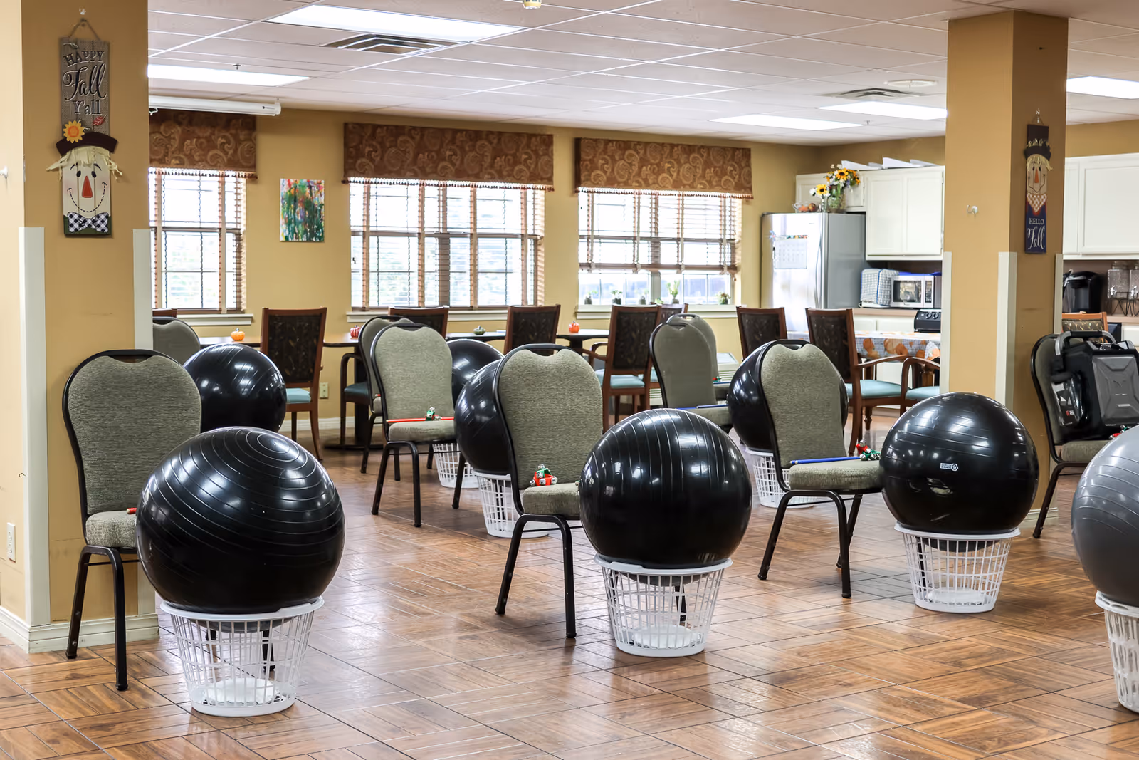 A spacious room with several chairs arranged in rows, each paired with a large black exercise ball resting on an upside-down white basket. The room has large windows with brown patterned valances, wooden flooring, and a kitchen area with white cabinets and appliances in the background. Fall-themed decorations hang on the walls.