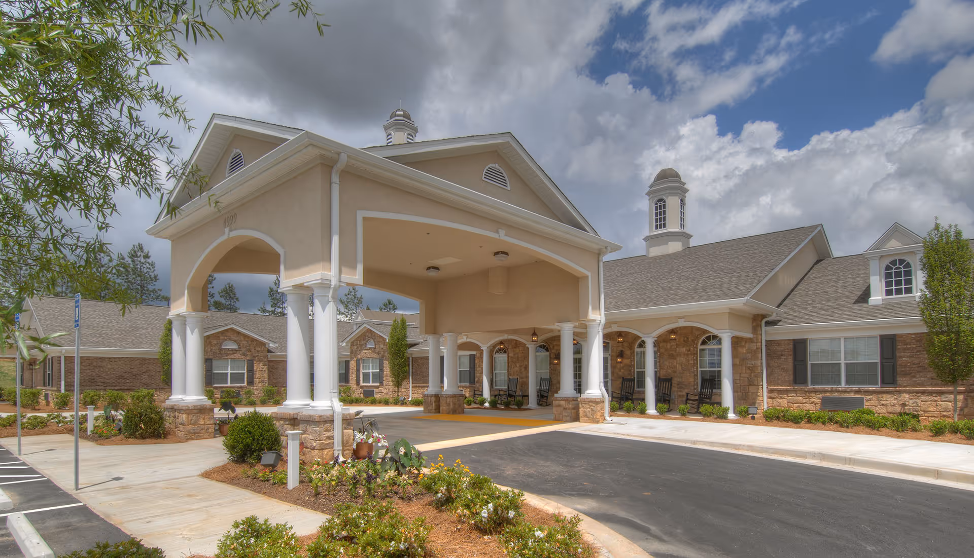 Front entrance of Benton House of Douglasville showing a covered porte-cochere with white columns, a porch with rocking chairs, and a landscaped driveway under a cloudy sky.