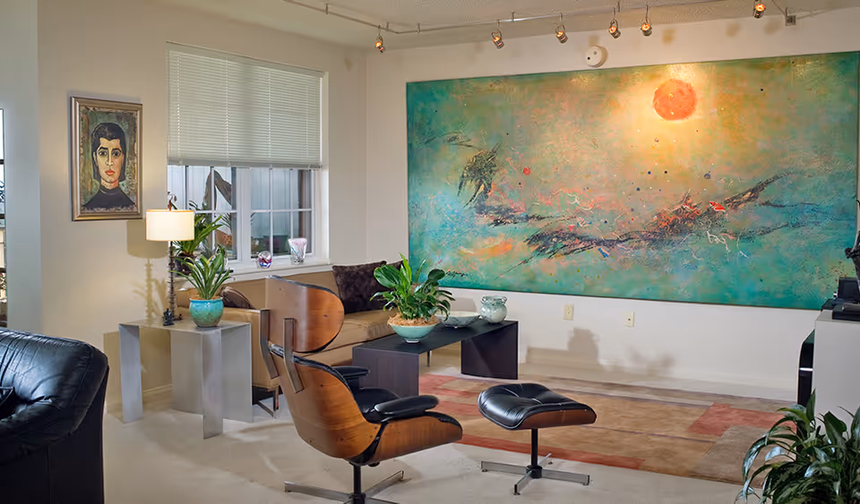 A cozy living room with a modern black leather lounge chair and ottoman, a beige sofa with dark cushions, a black coffee table with green plants, a large colorful abstract painting on the wall, a window with blinds, a side table with a lamp and a potted plant, and a framed portrait on the wall.