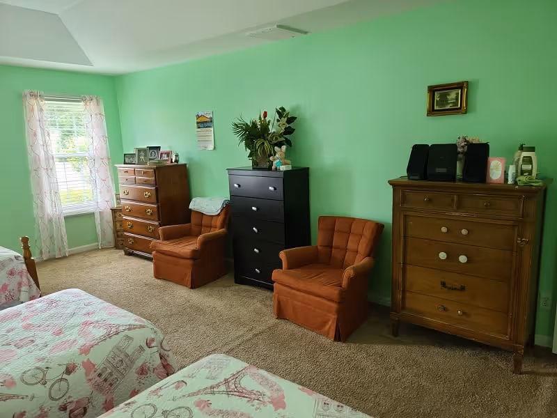 A bedroom with light green walls featuring two twin beds with pink and white patterned bedspreads. There are two brown cushioned armchairs placed between three wooden dressers of varying styles and colors. A window with sheer curtains allows natural light into the room, and decorative items such as framed photos, a calendar, and a vase with flowers are placed on the dressers.