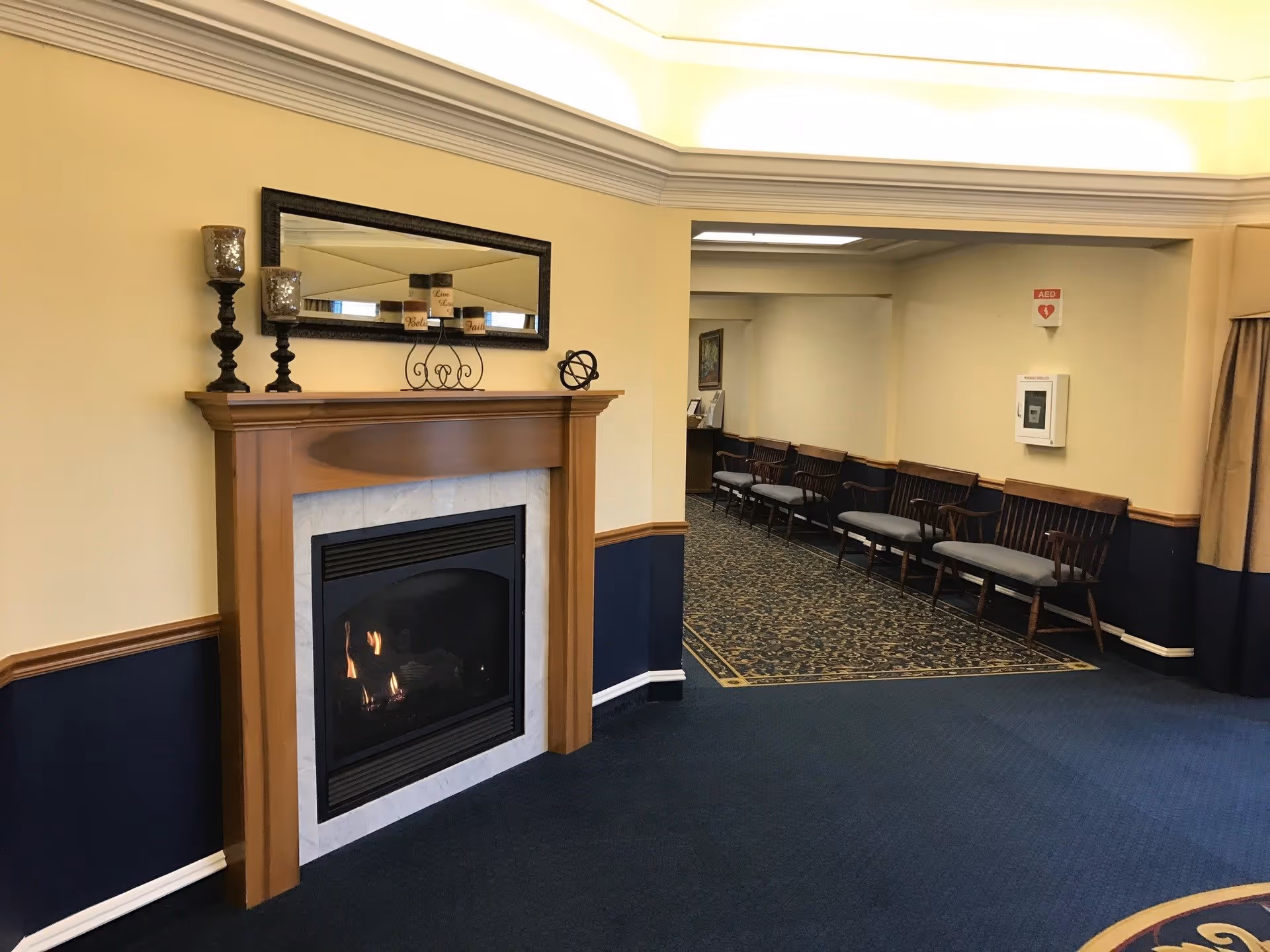 Interior view of a senior living facility lounge area featuring a lit fireplace with a wooden mantle, decorative candles, and a mirror above it. To the right, there is a hallway with several wooden chairs lined up against the wall and an AED mounted on the wall.